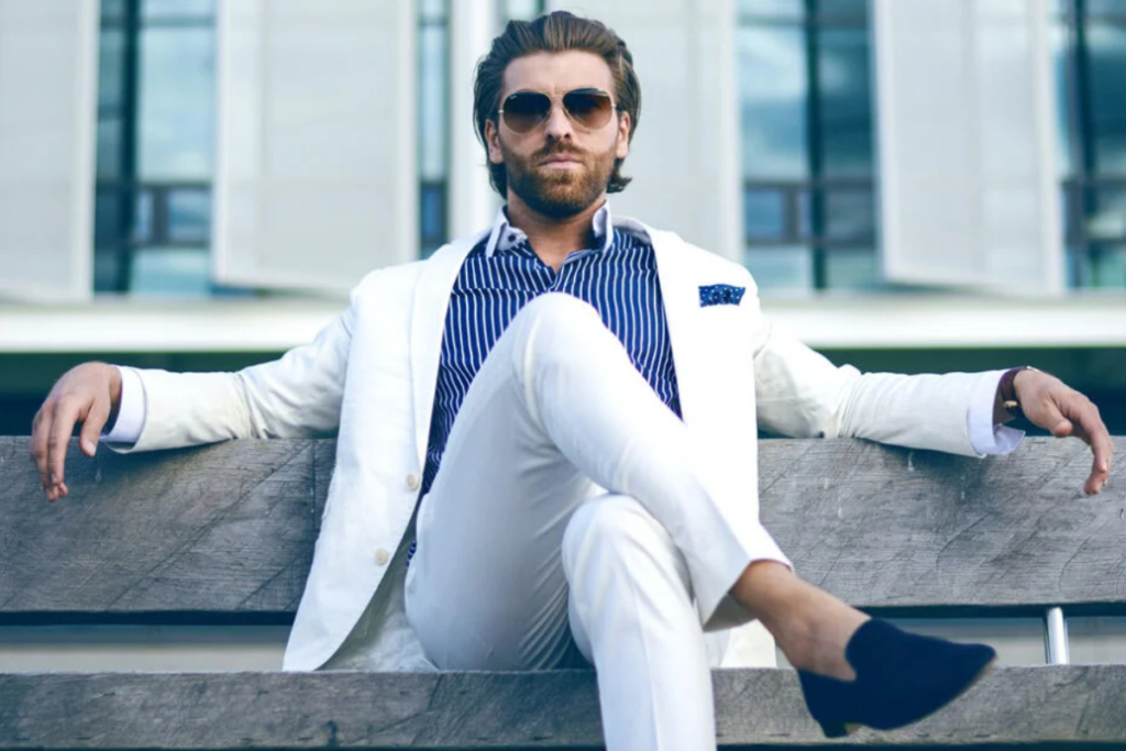 A stylish man in a white suit and sunglasses sitting confidently on a bench, with a modern building in the background.