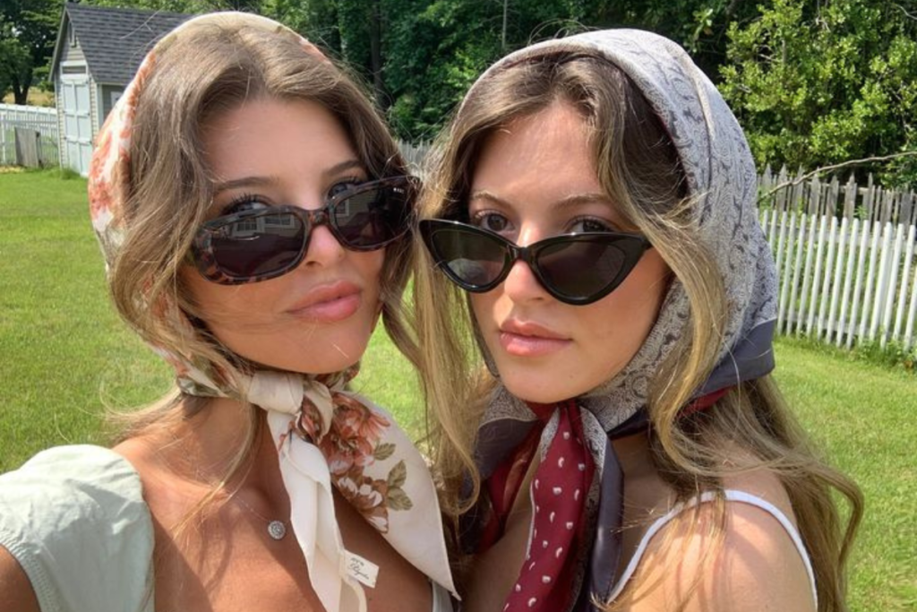 Two women wearing sunglasses and stylish headscarves, posing outdoors with a scenic green background and white fence.