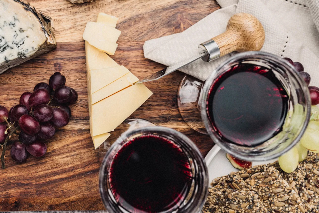 Overhead view of a wine and cheese spread with red wine, assorted cheeses, grapes, and seeded crackers on a rustic wooden board.