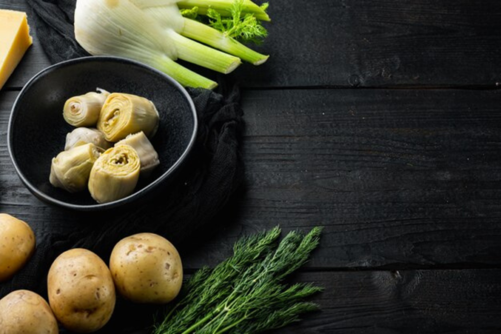 Fresh ingredients on a dark wooden surface, including fennel, artichoke hearts in a bowl, potatoes, and dill, ready for cooking preparation.