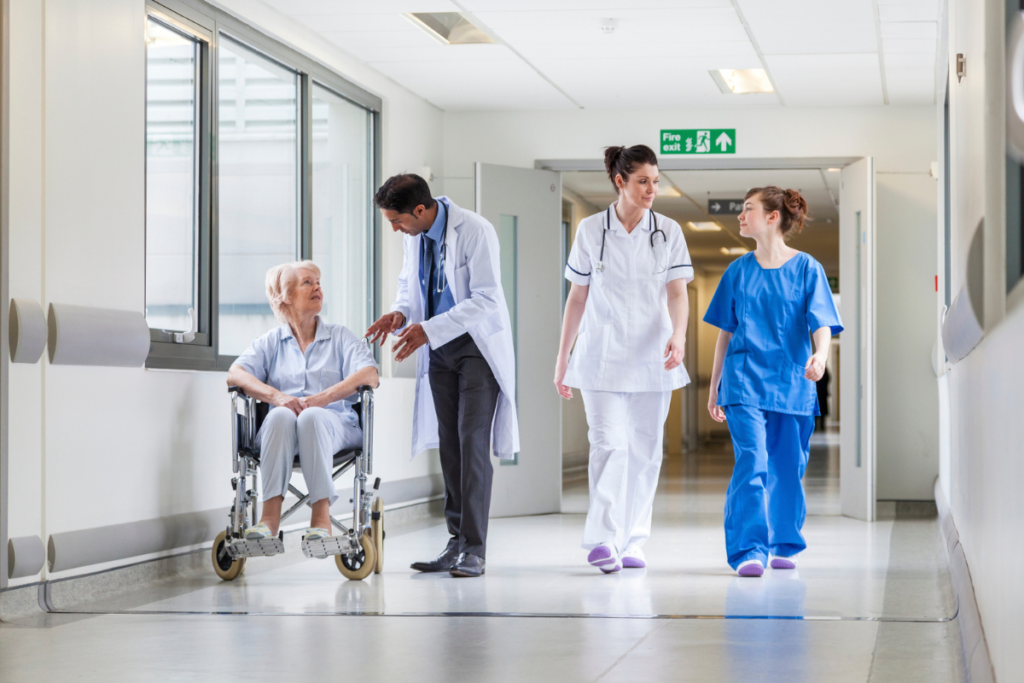 Doctor and nurses assist an elderly patient in a wheelchair down a hospital corridor.