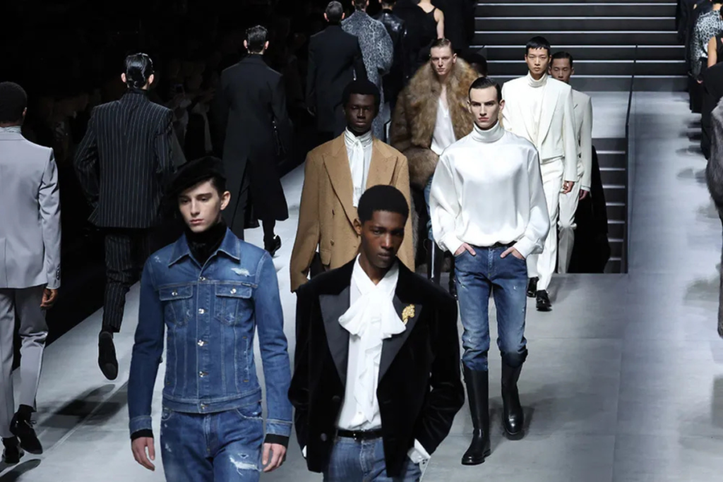 Runway fashion show with male models in diverse styles, from denim to tailored suits and fur coats.