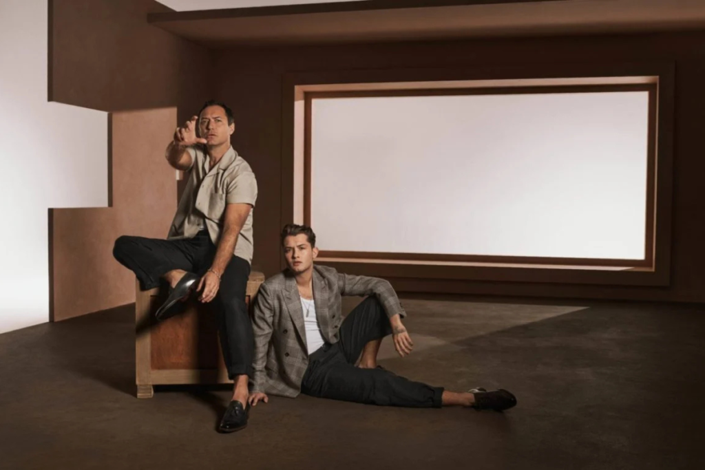 Two men pose in a minimalist room with neutral tones, combining relaxed and formal fashion styles.