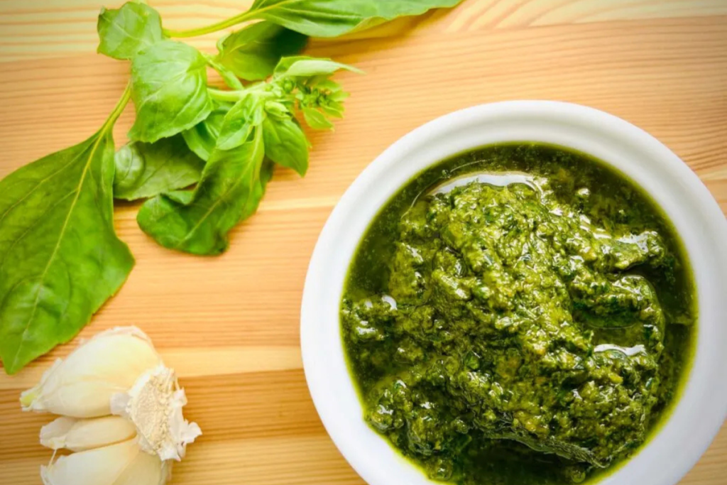 Bowl of fresh pesto with basil leaves and garlic cloves on a wooden surface, showcasing ingredients for a classic Italian sauce recipe.