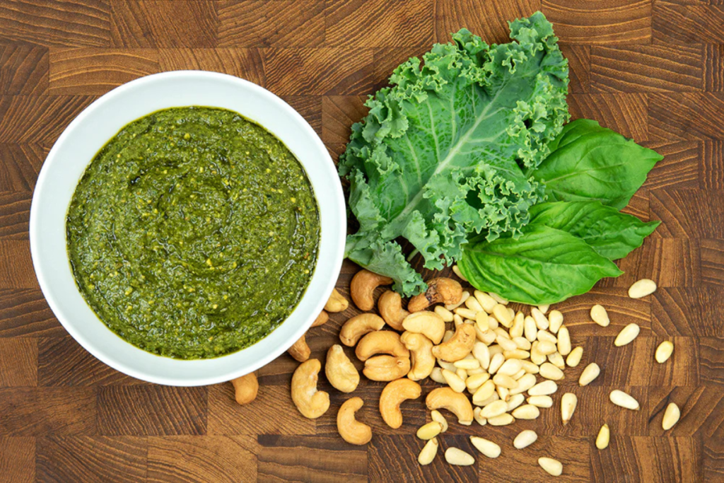 Bowl of vibrant green pesto with fresh kale, basil leaves, cashews, and pine nuts on a wooden background, ideal for a nutritious recipe.