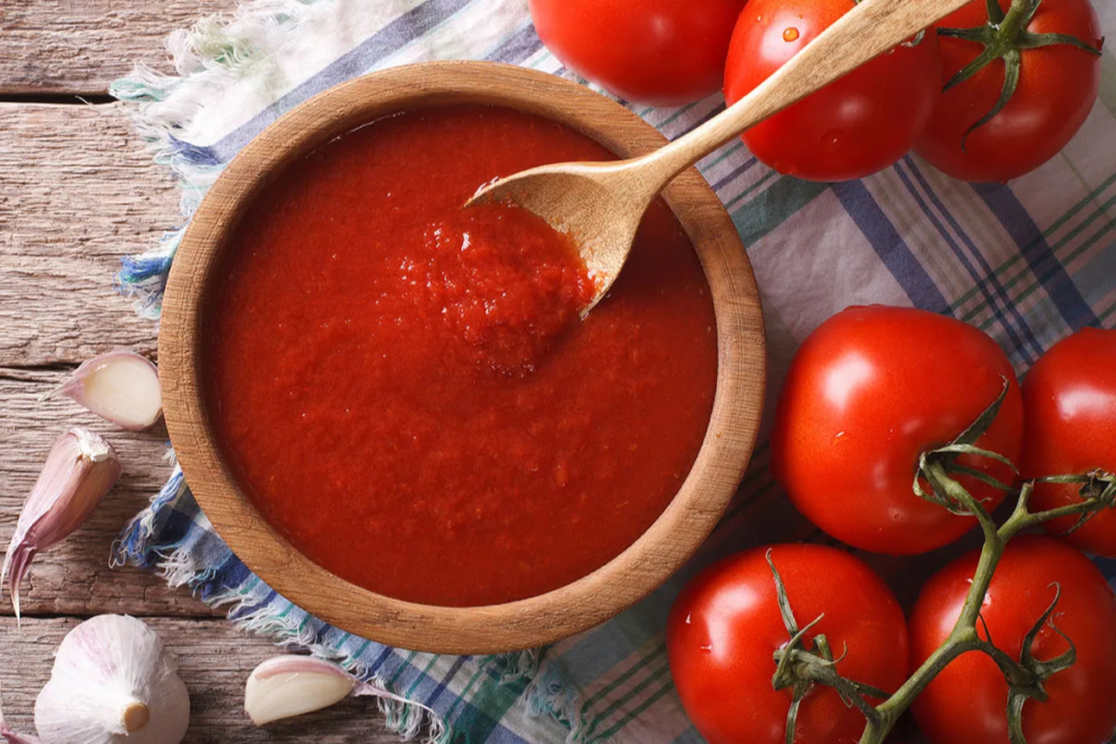 Fresh tomato sauce in a wooden bowl with a spoon, surrounded by ripe tomatoes on the vine, garlic cloves, and a rustic cloth on wooden table.