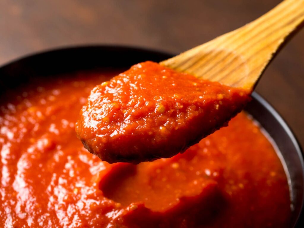 Close-up of a wooden spoon lifting a thick, rich tomato sauce, showcasing its smooth texture and vibrant red color in a dark bowl.